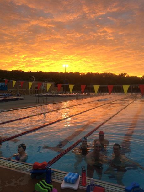 A group of swimmers smiling in an Ann Arbor outdoor pool with a golden sunset in the sky behind them