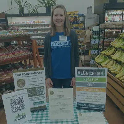 A woman wearing a VegMichigan tshirt stands behind a table at a grocery store with informational flyers.