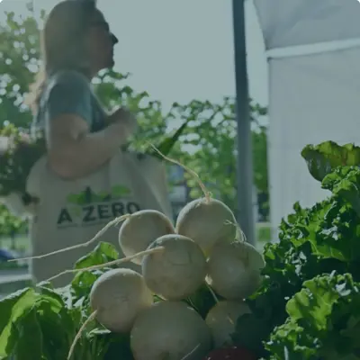 A person holding a reusable bag with the A2ZERO logo on it stands behind a farmers market table with piles of turnips and radishes on top.