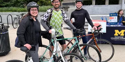 Bike to Wherever Day event participants are featured with their bikes, a man, woman and male teen, alongside a University of Michigan information table
