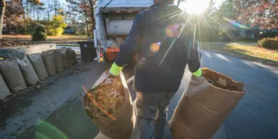 A municipal worker carries a paper yard waste bag full of leaves one in each hand toward a collection truck parked on a residential street