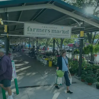 People carrying full grocery bags walk beneath a pavilion under a sign that says "Ann Arbor Farmers Market"