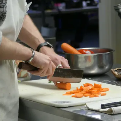 A person wearing jewelry and an apron chops a carrot on a white cutting board.
