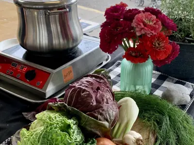 Table with electric stove, pan, and a variety of vegetables and flowers, including cabbage, garlic, fennel, and onions.