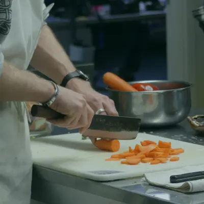 A person wearing jewelry and an apron chops a carrot on a white cutting board.